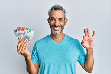 Middle age grey-haired man holding australian dollars doing ok sign with fingers, smiling friendly...