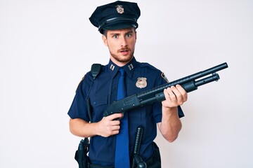Young caucasian man wearing police uniform holding shotgun in shock face, looking skeptical and sarcastic, surprised with open mouth