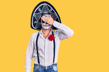 Man wearing day of the dead costume over background covering eyes with hand, looking serious and sad. sightless, hiding and rejection concept