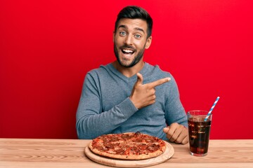 Handsome hispanic man eating tasty pepperoni pizza smiling cheerful pointing with hand and finger up to the side