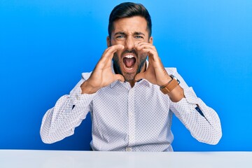 Handsome hispanic man wearing business clothes sitting on the table shouting angry out loud with hands over mouth