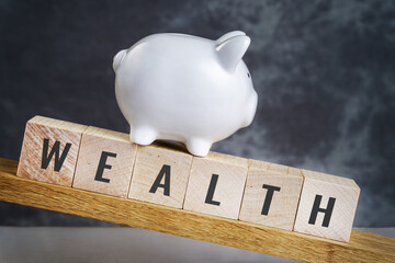 Word wealth spelled in letters on wooden block under the piggy bank tilted down against dark...
