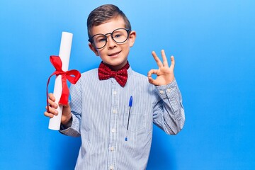 Cute blond kid wearing nerd bow tie and glasses holding diploma doing ok sign with fingers, smiling...