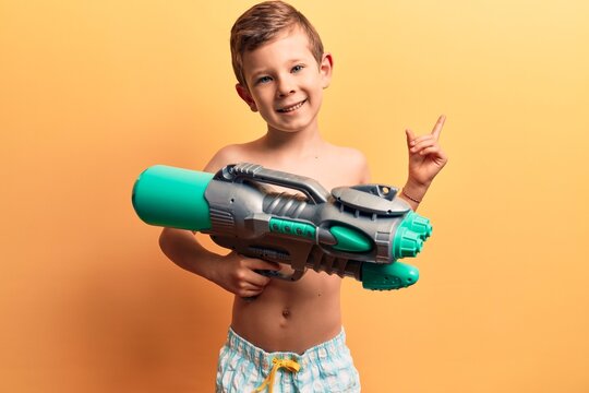 Cute blond kid wearing swimwear holding water gun smiling happy pointing with hand and finger to the side