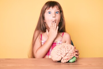 Little caucasian kid girl with long hair holding brain as mental health concept covering mouth with...