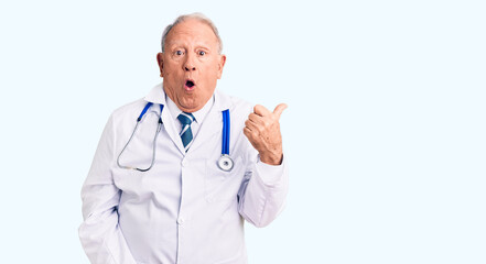 Senior handsome grey-haired man wearing doctor coat and stethoscope surprised pointing with hand...