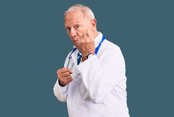 Senior handsome grey-haired man wearing doctor coat and stethoscope ready to fight with fist...