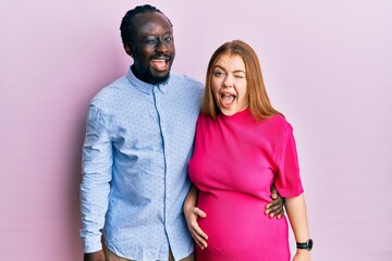 Young interracial couple expecting a baby, touching pregnant belly winking looking at the camera...