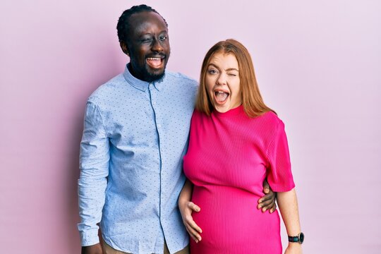 Young interracial couple expecting a baby, touching pregnant belly winking looking at the camera with sexy expression, cheerful and happy face.