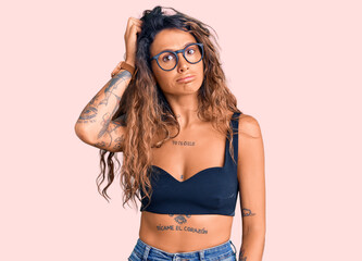 Young hispanic woman with tattoo wearing casual clothes and glasses confuse and wonder about...