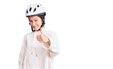 Beautiful brunette young woman wearing bike helmet and sporty clothes doing happy thumbs up gesture with hand. approving expression looking at the camera showing success.