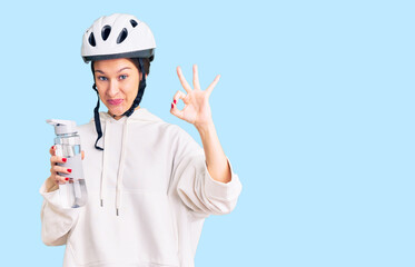 Beautiful brunette young woman wearing bike helmet and holding water bottle doing ok sign with fingers, smiling friendly gesturing excellent symbol