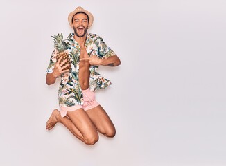 Young handsome hispanic man on vacation wearing swimwear, floral shirt and hat smiling happy. Jumping with smile on face holding pineapple over isolated white background
