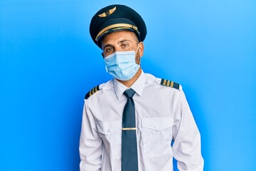 Handsome man with beard wearing airplane pilot uniform wearing safety mask with serious expression on face. simple and natural looking at the camera.