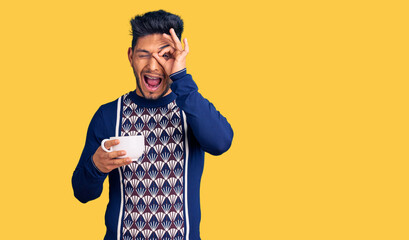 Handsome latin american young man drinking a cup of coffee smiling happy doing ok sign with hand on eye looking through fingers