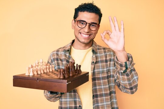 Young handsome hispanic man holding chess board wearing glasses doing ok sign with fingers, smiling friendly gesturing excellent symbol