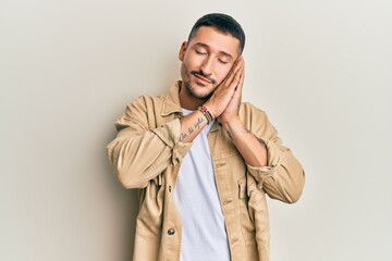 Handsome man with tattoos wearing casual brown denim jacket sleeping tired dreaming and posing with hands together while smiling with closed eyes.