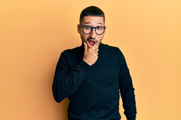 Handsome man with tattoos wearing turtleneck sweater and glasses looking fascinated with disbelief, surprise and amazed expression with hands on chin