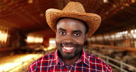 Portrait of handsome cheerful man in hat and motley shirt standing in stable of sheep farm rising...