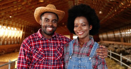 Portrait of happy African American young couple of farmers standing together in hugs in sheep farm shed. Beautiful woman and handsome man hugging in stable. Dolly shot. Male and female joyful workers.