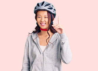 Young beautiful chinese girl wearing bike helmet pointing finger up with successful idea. exited and happy. number one.