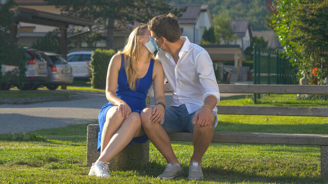 PORTRAIT: Cuddling Couple Wearing Facemasks Kisses While Sitting On A Park Bench