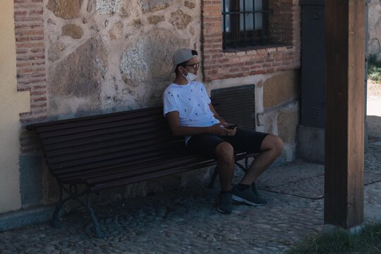 young man sitting on a bench with a health mask waiting and using his mobile phone to communicate via messages or social networks