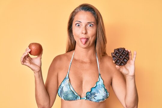 Middle age hispanic woman holding apple and donut sticking tongue out happy with funny expression.