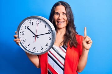 Young beautiful brunette woman doing countdown holding big clock over blue background smiling with an idea or question pointing finger with happy face, number one