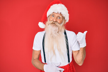 Fototapeta na wymiar Old senior man with grey hair and long beard wearing white t-shirt and santa claus costume smiling with happy face looking and pointing to the side with thumb up.