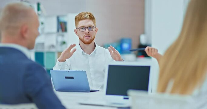 Young man with red hair is sitting in negotiations with partners and talking, a business meeting in a business center. Business colleagues negotiations, laptops on the table. 4k, ProRes