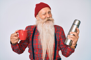 Old senior man with grey hair and long beard holding cup of coffee and thermo winking looking at...
