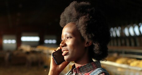Young African American woman talking on cellphone and working in farm stable. Female farmer speaking on mobile phone in shed. Shepherd having call and conversation on telephone. Farming concept.