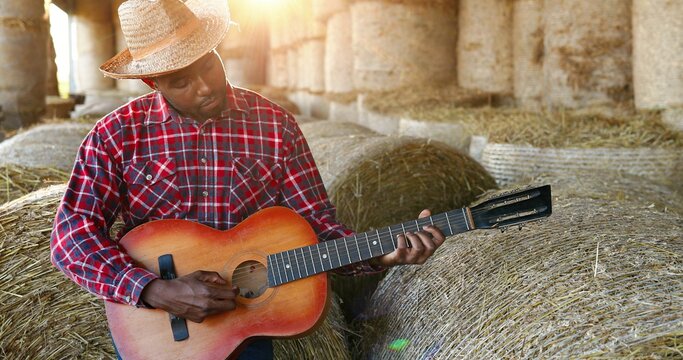 African American young handsome man sitting on hay stocks in shed and playing music on guitar. Male farmer singing and play on musical instrument song. Countryside concept. Village.