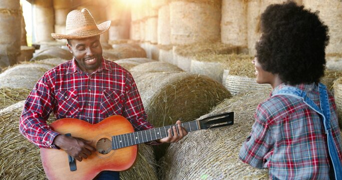 African American young man farmer in hat sitting on hay stocks and playing on guitar for pretty woman. Couple of male and female farmers in shed. Romantic evening in village. Guy singing love song. - Powered by Adobe