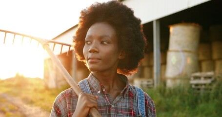 African American woman with curly hair walking in field early in morning with pitchfork. Farming lifestyle. female farmer holding fork and going to work outdoor. Girl in village.