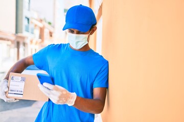 Fototapeta na wymiar African delivery man wearing courier uniform outdoors wearing coronavirus safety mask holding cardboard parcel using smartphone