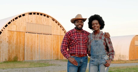 Portrait of happy African American young couple of farmers hugging outdoors at shed on background and smiling to camera in village. Man and woman with smiles in hugs. Farming concept. Embracing.