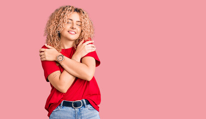 Young blonde woman with curly hair wearing casual red tshirt hugging oneself happy and positive,...