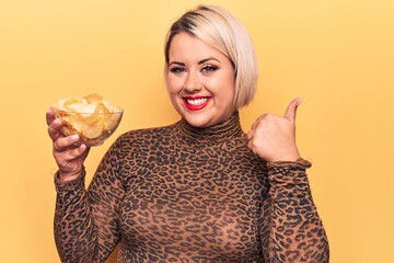 Beautiful blonde plus size woman holding bowl with potatoes chips over yellow background smiling...
