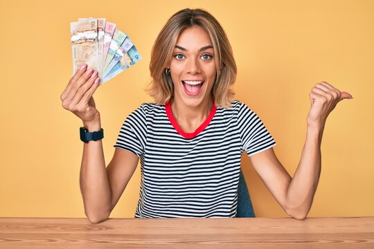 Beautiful caucasian woman holding canadian dollars pointing thumb up to the side smiling happy with open mouth