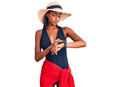 Young african american woman wearing swimsuit and summer hat checking the time on wrist watch, relaxed and confident