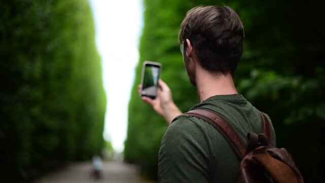 Young Handsome Man Traveler with Backpack on His Back Makes a Video on a Smartphone in a Park.Tourist Shooting a Stories Clip on for Social Media by Phone in Green tree labyrinth.