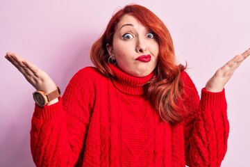 Young beautiful redhead woman wearing red casual turtleneck sweater over pink background clueless and confused with open arms, no idea and doubtful face.