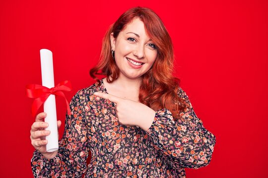 Young beautiful redhead woman holding graduated degree diploma over red background smiling happy pointing with hand and finger