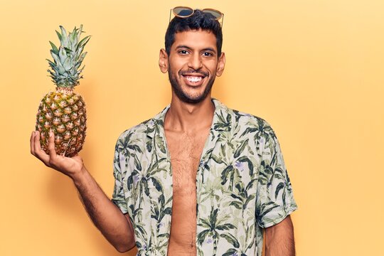 Young latin man wearing summer shirt holding pineapple looking positive and happy standing and smiling with a confident smile showing teeth