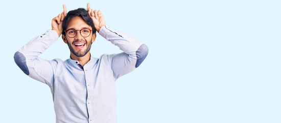 Handsome hispanic man wearing business shirt and glasses doing funny gesture with finger over head as bull horns