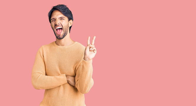 Handsome hispanic man wearing casual sweater smiling with happy face winking at the camera doing victory sign. number two.