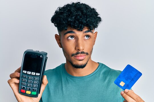 Young arab handsome man holding dataphone and credit card smiling looking to the side and staring away thinking.