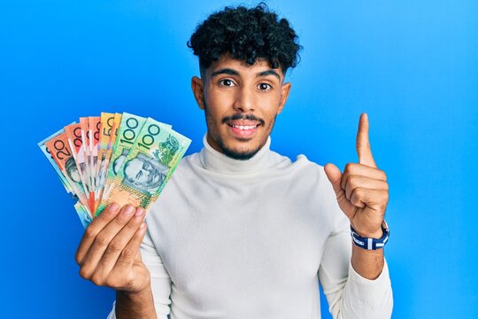 Young arab handsome man holding australian dollars smiling with an idea or question pointing finger with happy face, number one